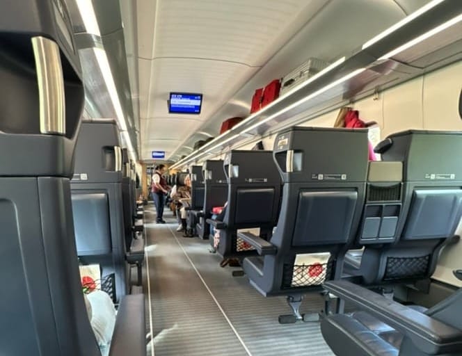 A picture taken from the seat of a Deutsche-Bahn ICE train: a corridor of comfortable large train chairs. Down the aisle, an attendant is serving a passenger.